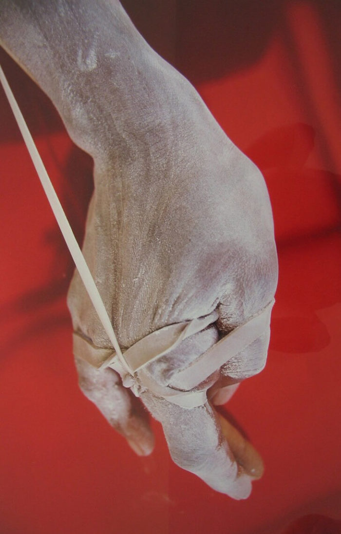 Myriam Mihindou, incarnation, série de photographies couleur où l'on voit une main enserrée dans de gros élastiques, sur fond rouge. photographie contemporaine Maroc