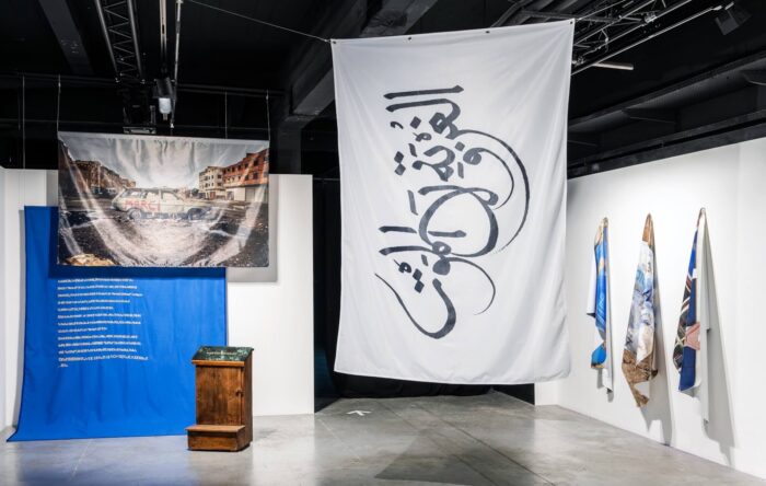 à gauche: Mohammed Laouli Merci Renault, 2026 Installation, broderie en fil dorée sur toile de travail bleu, 260 x 200cm, photographie imprimée sur tissu, 200 x 130 cm, prie-dieu en bois brut, plaque en marbre gravée et dorée, 47 x 22 cm. Courtesy de l’artiste et de l’adagp, Paris, 2026. à droite: Katrin Ströbel & Mohammed Laouli frontières fluides - L’exil ou la mort, 2017 Drapeau, 150 x 100cm Courtesy des artistes et de l’adagp, Paris, 2026. © La Condition Publique & Le Cube - independent art room photo: Frédéric IOVINO